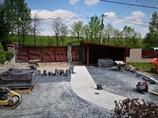 Układanie kostki brukowej: częściowo ułożona ścieżka z jasnej kostki, obok palety z ciemniejszą kostką, zagęszczarka i narzędzia na posesji z czerwonym ogrodzeniem.