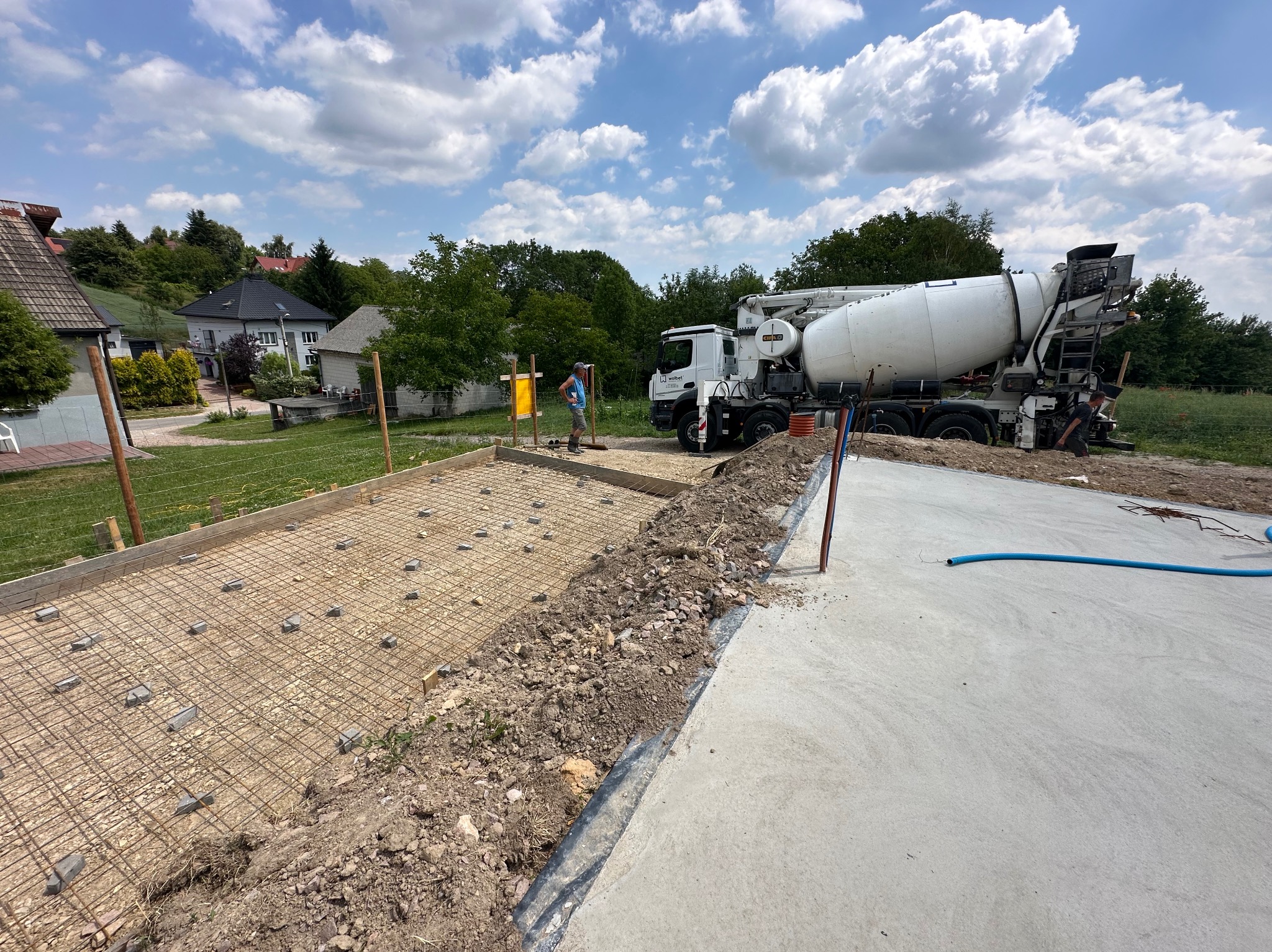 Przygotowanie zbrojenia pod wylewkę fundamentu z użyciem siatki stalowej i dystansów, z betoniarką w tle, na tle zabudowy jednorodzinnej w słoneczny dzień.