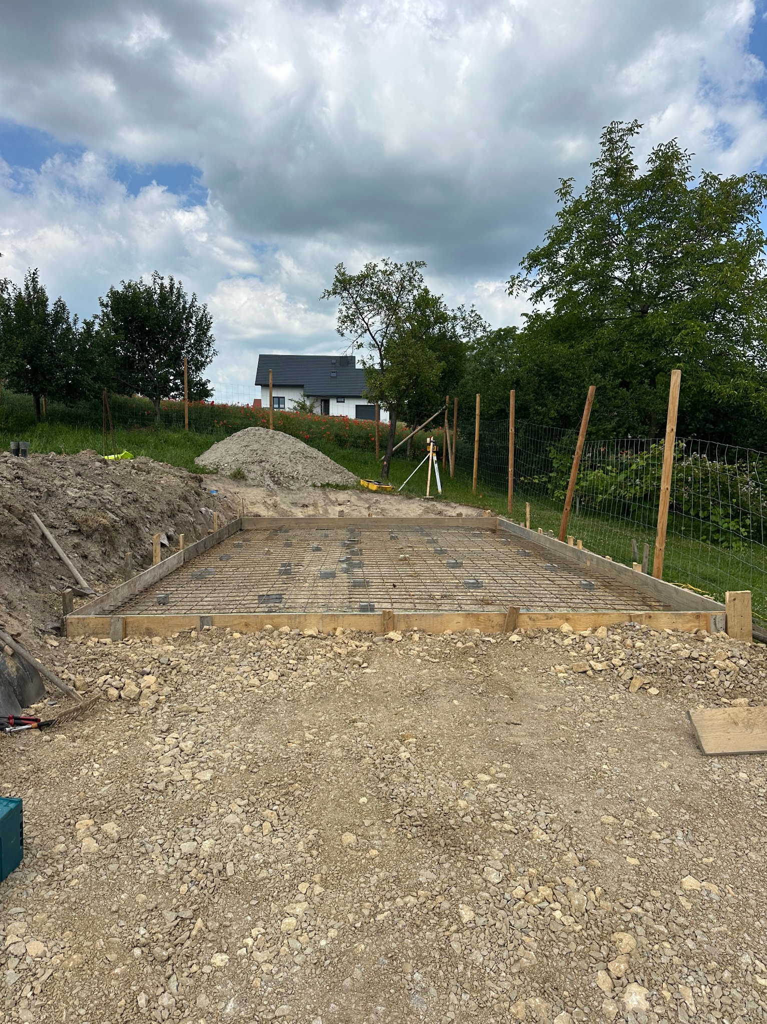 Zbrojenie fundamentu pod budowę w Jerzmanowicach. Widoczna siatka zbrojeniowa w drewnianym szalunku, przygotowana do zalania betonem. W tle dom i drzewa.