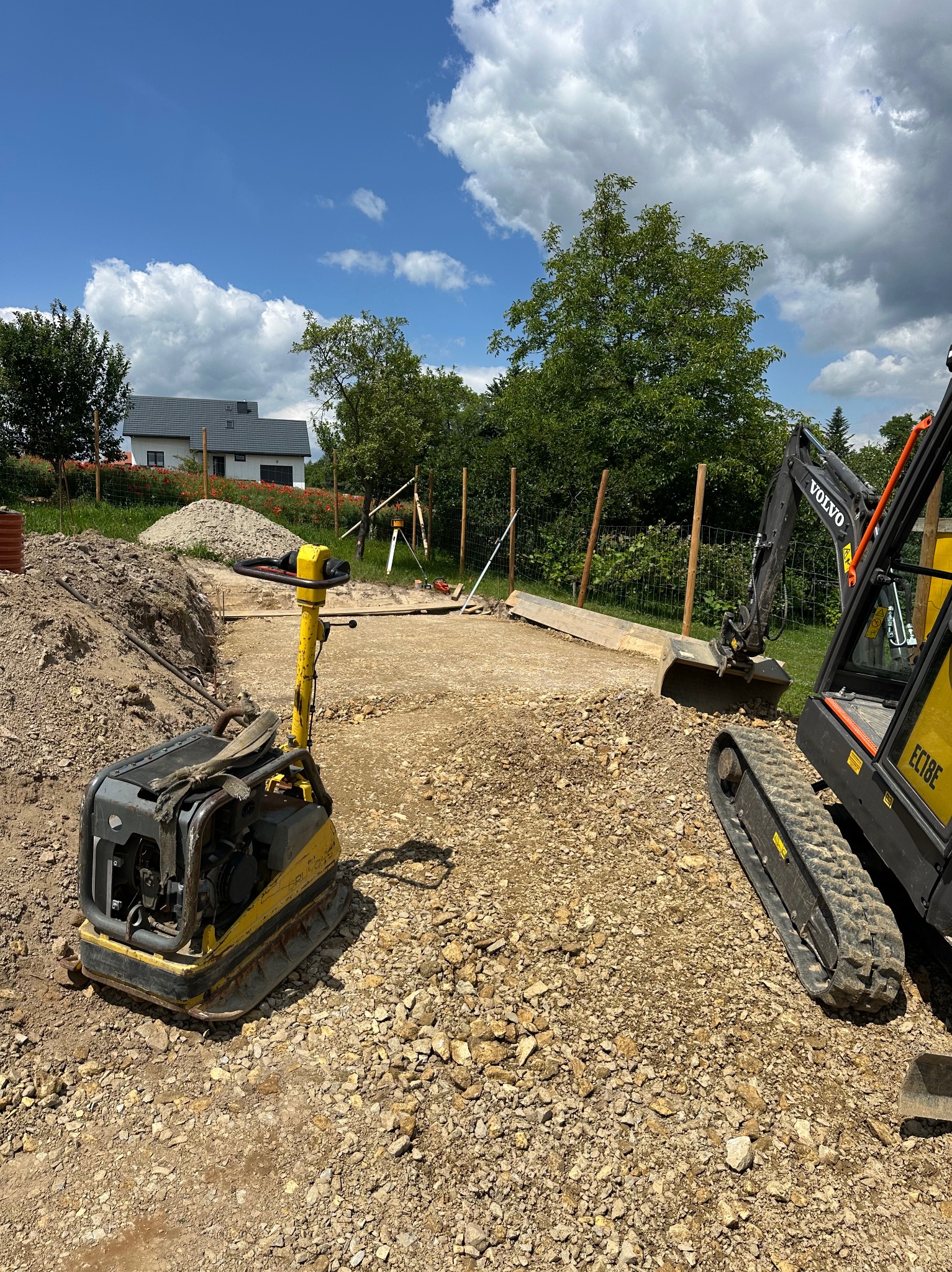 Żółta ubijarka i minikoparka Volvo na placu budowy z przygotowanym terenem pod fundament. W tle dom z szarą dachówką i błękitne niebo z obłokami.
