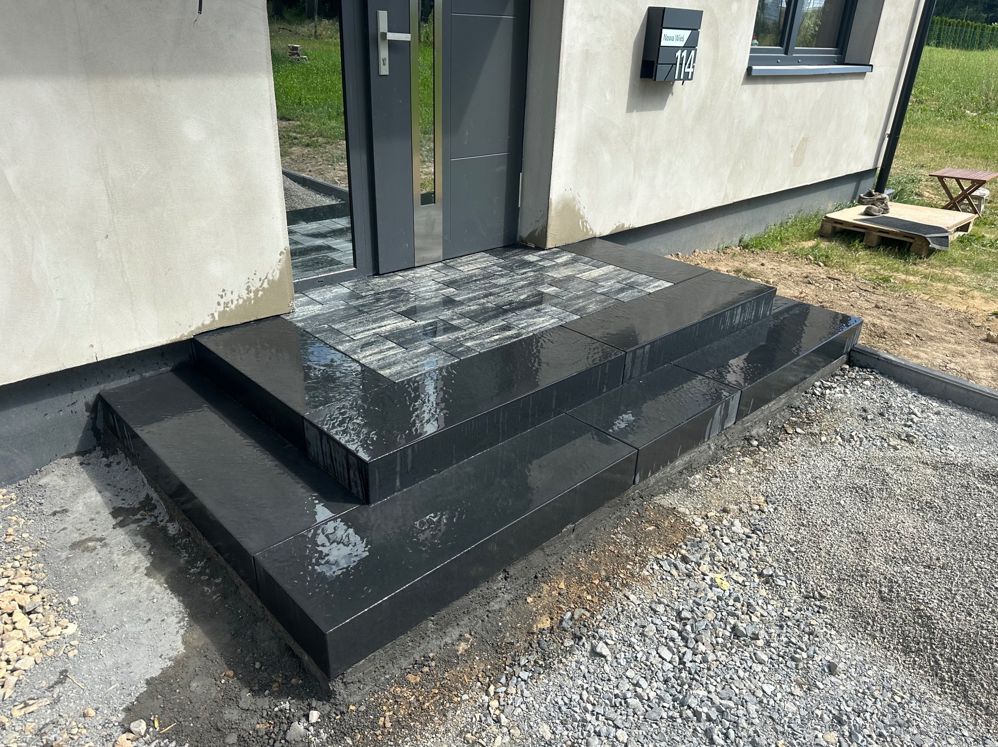 Nowe, czarne schody z kostki brukowej prowadzące do drzwi wejściowych domu, wykończone szarą kostką na podeście. Widoczne niedokończone otoczenie.