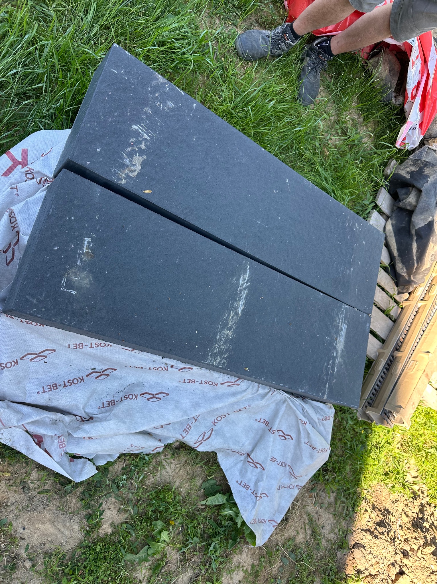 Dwa czarne bloki styropianowe na folii z logo Kost-Bet, leżące na trawie. Widoczne fragmenty obuwia i narzędzi. Przygotowania do izolacji fundamentów.