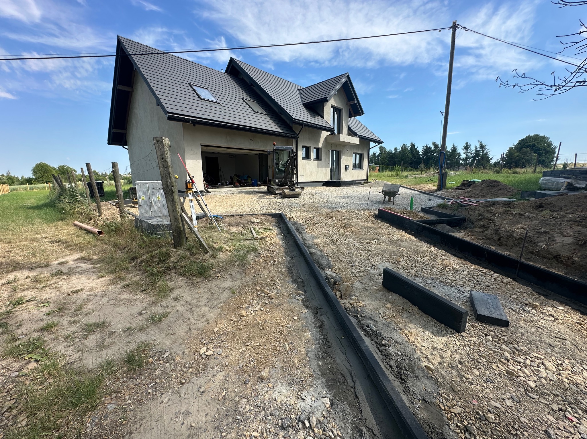 Budowa domu jednorodzinnego: przygotowanie terenu pod kostkę brukową, widoczne obrzeża i mini koparka na żwirowym podłożu, dom w tle.