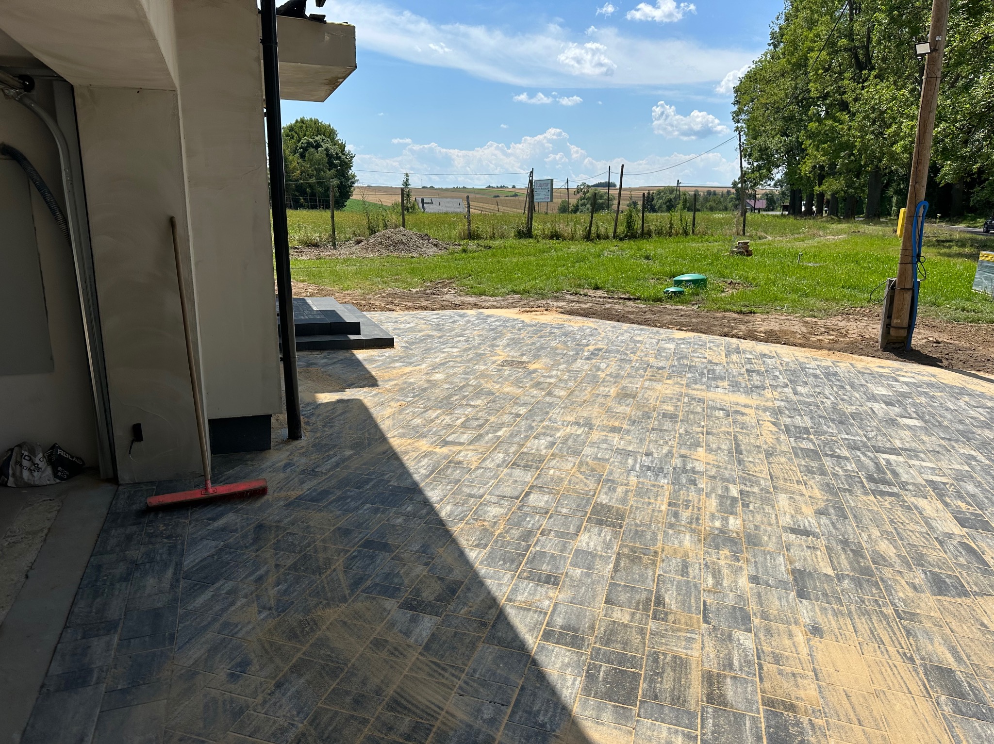 Nowo ułożona, szara kostka brukowa na podjeździe, z widocznym fragmentem budynku i otwartym garażem. W tle zielony krajobraz i błękitne niebo.