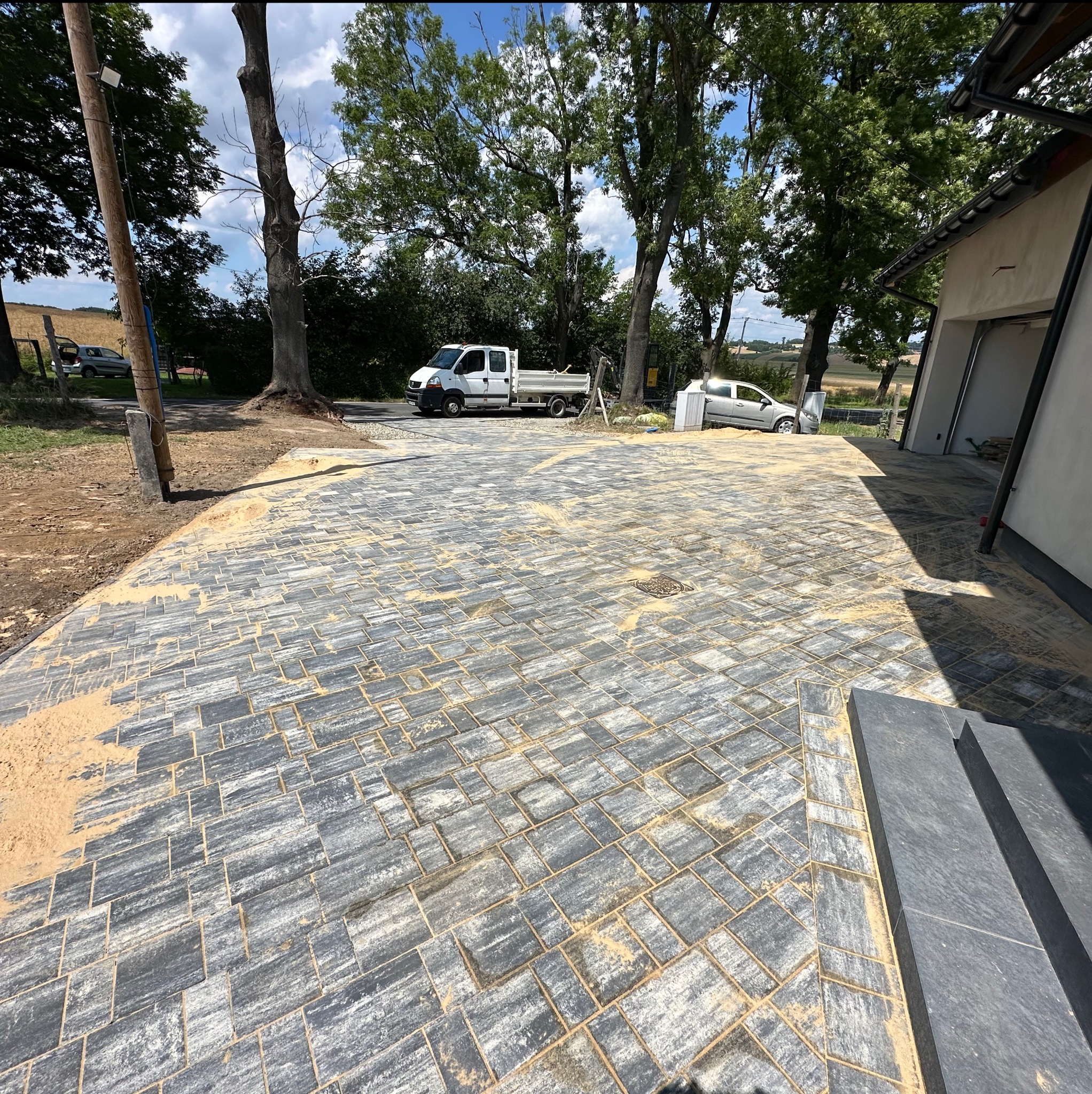 Świeżo ułożona, szara kostka brukowa z piaskiem na podjeździe, widok z góry. W tle drzewa, biały samochód i budynek z garażem.