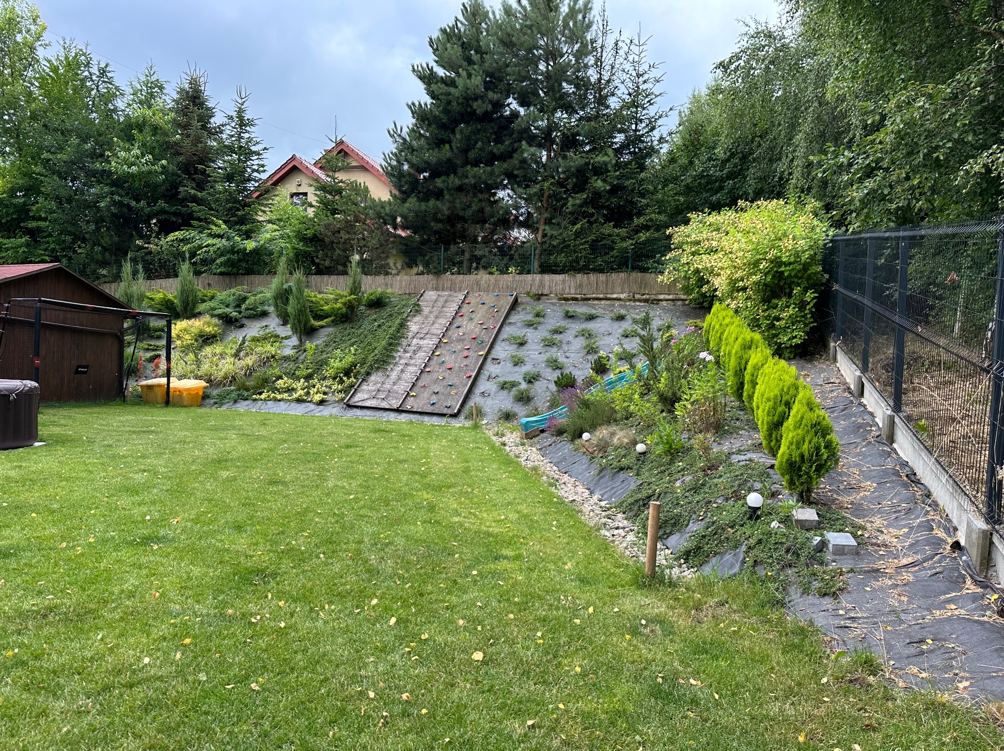 Zielony ogród ze ścianką wspinaczkową na wzgórzu, z równo przystrzyżonym trawnikiem i dekoracyjnymi krzewami wzdłuż ścieżki z siatki, w tle dom.