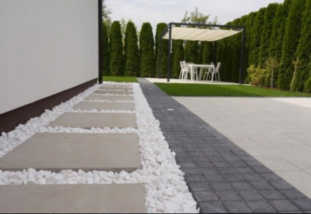 Nowoczesny ogród z ścieżką z płyt otoczonych białym grysem, prowadzącą do pergoli z meblami ogrodowymi i zadbanym trawnikiem, otoczony wysokimi tujami.