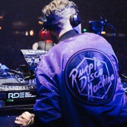 BARTŁOMIEJ SZOPA SHORE STUDIO - DJ w słuchawkach, odwrócony tyłem, w fioletowej kurtce z logo 'Purple Disco Machine' obsługuje sprzęt DJ-ski podczas występu na evencie. W tle widoczny sprzęt i oświetlenie sceniczne.