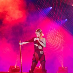 BARTŁOMIEJ SZOPA SHORE STUDIO - Piosenkarka z mikrofonem na scenie w kostiumie z wycięciami, oświetlona czerwonymi i niebieskimi światłami, trzymająca brokatową laskę.