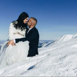 Para młoda w zimowej scenerii górskiej, delikatny pocałunek na tle błękitnego nieba. Panna młoda w białej sukni i futrze, pan młody w granatowym garniturze.