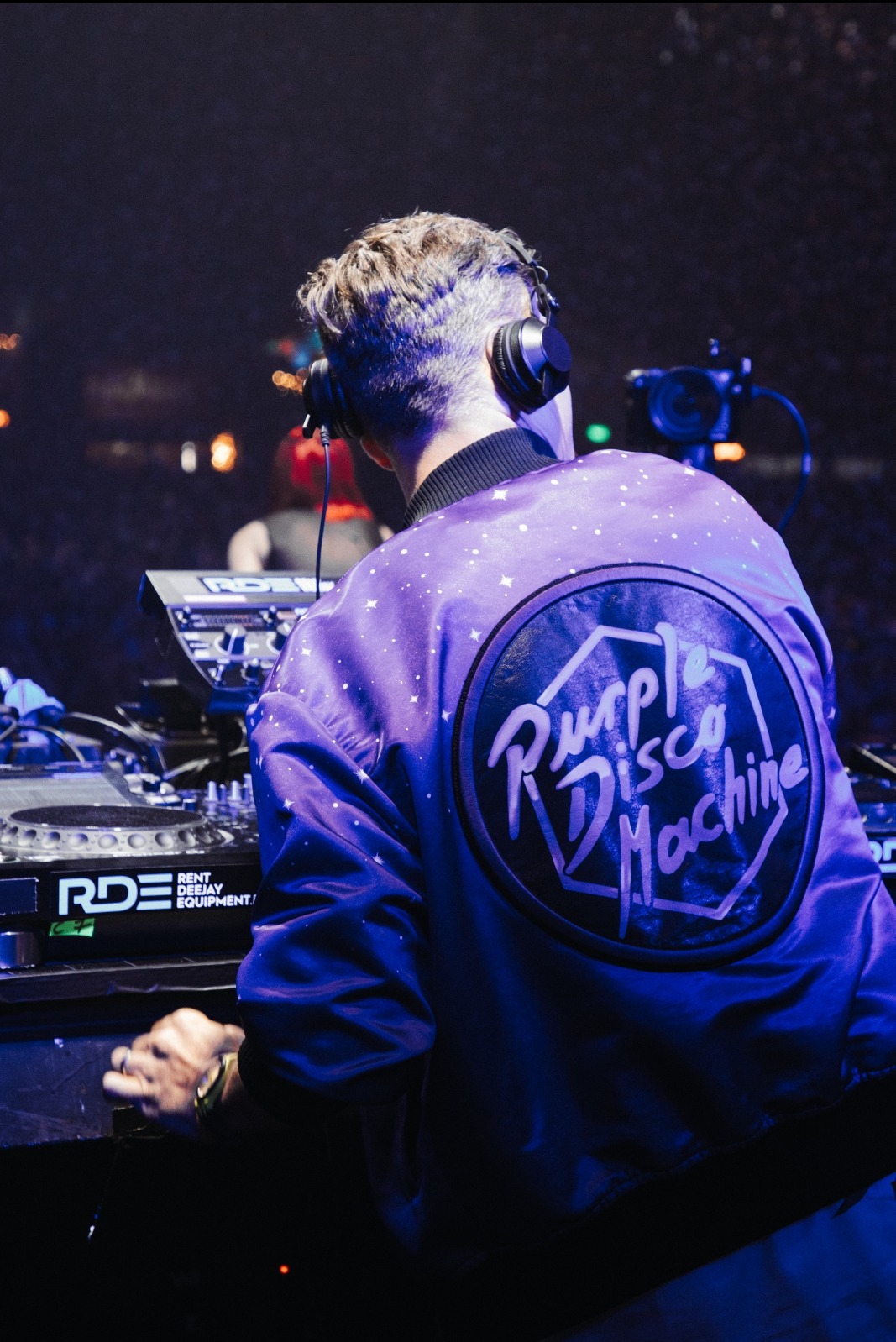 DJ w słuchawkach, odwrócony tyłem, w fioletowej kurtce z logo 'Purple Disco Machine' obsługuje sprzęt DJ-ski podczas występu na evencie. W tle widoczny sprzęt i oświetlenie sceniczne.