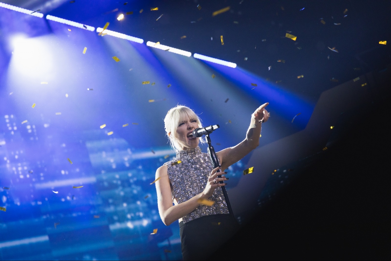 Piosenkarka w błyszczącej bluzce śpiewa do mikrofonu na scenie w świetle reflektorów, podczas gdy z góry opadają konfetti.
