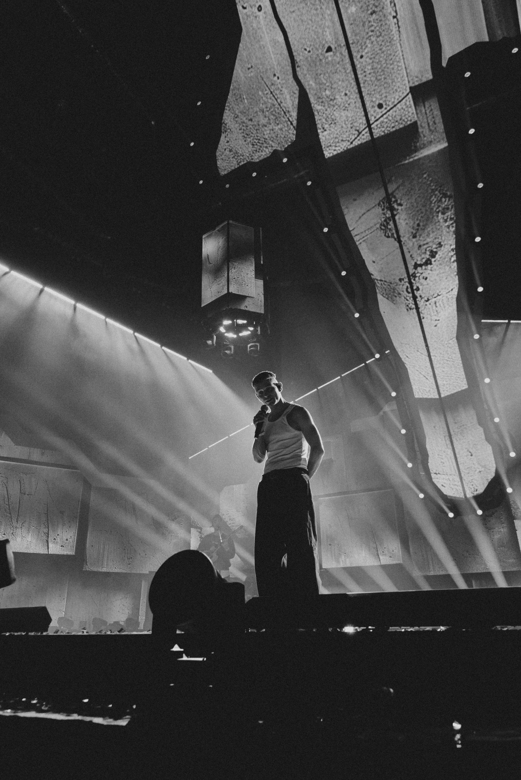 Czarno-białe zdjęcie piosenkarza na scenie podczas koncertu, oświetlonego reflektorami. W tle dekoracje i elementy scenografii.