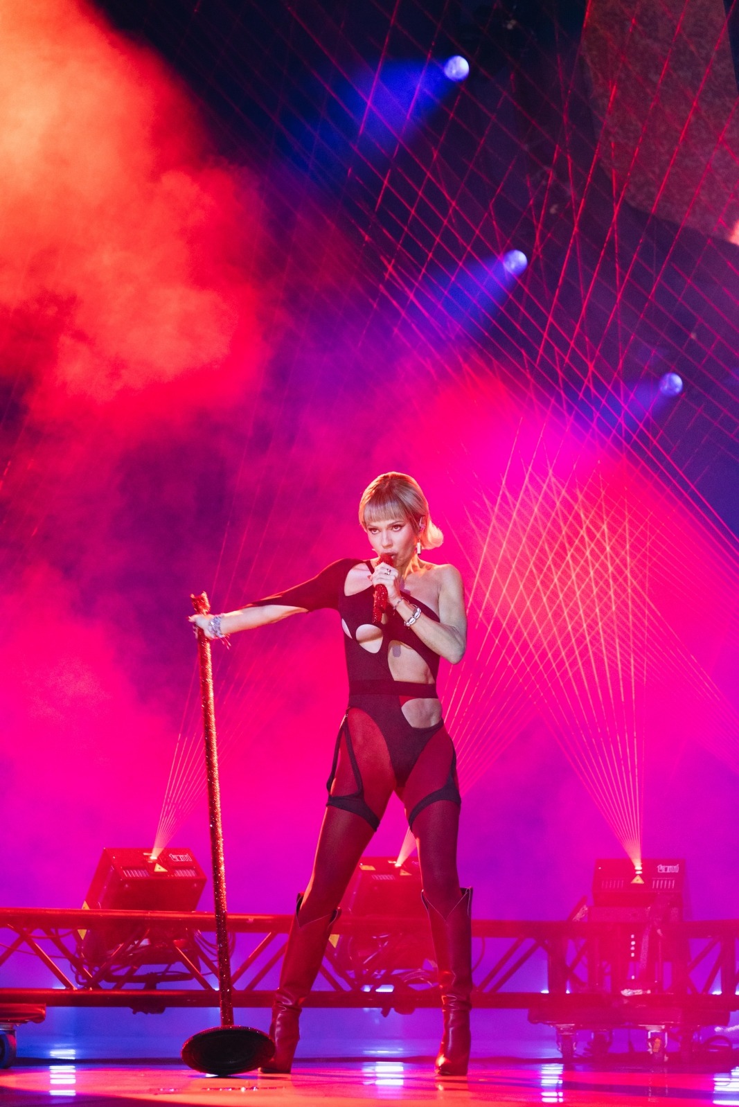 Piosenkarka z mikrofonem na scenie w kostiumie z wycięciami, oświetlona czerwonymi i niebieskimi światłami, trzymająca brokatową laskę.