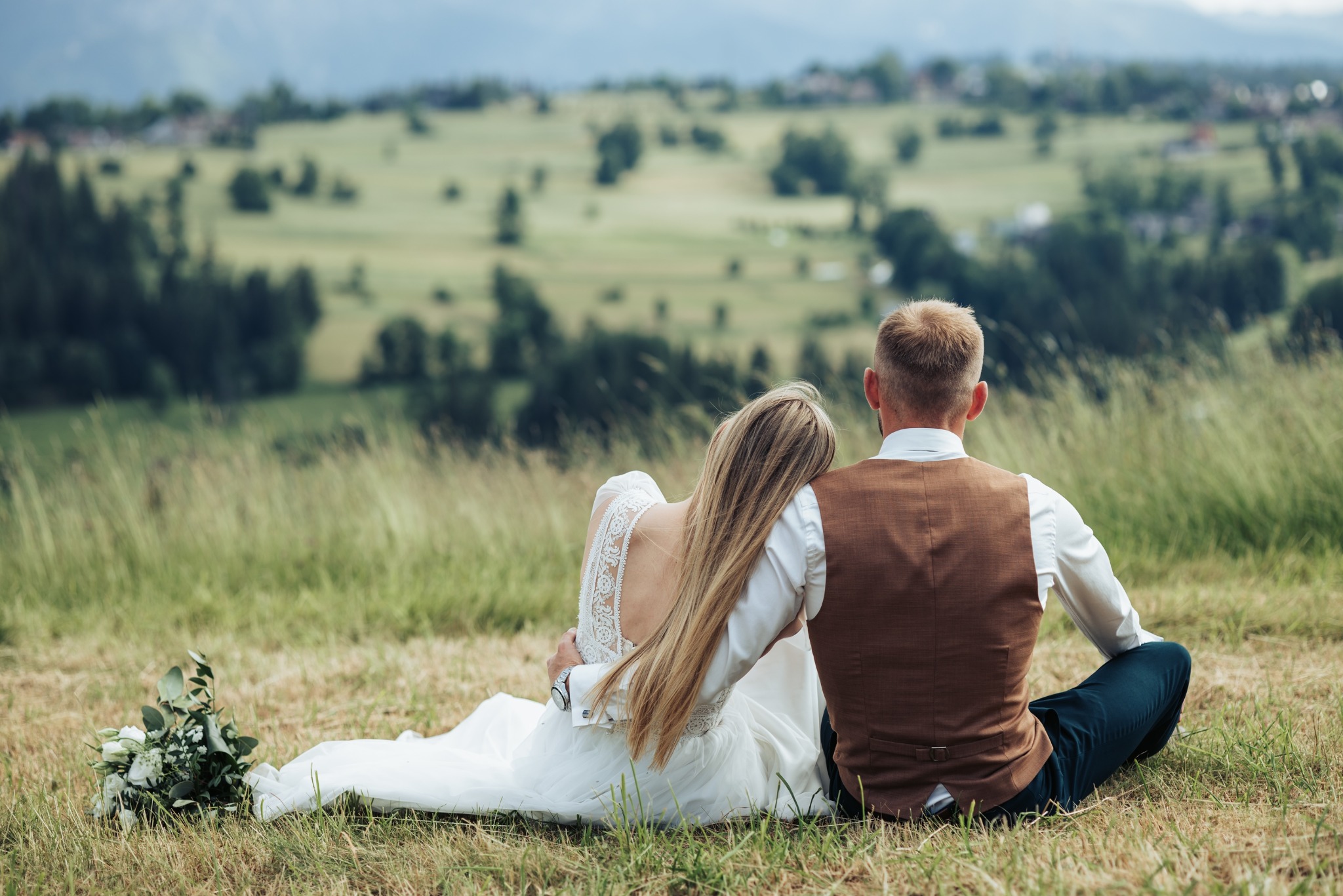 Para młoda siedzi na trawie, obejmując się, z widokiem na pagórkowaty krajobraz. Panna młoda w sukni z koronkowymi plecami, pan młody w brązowej kamizelce.