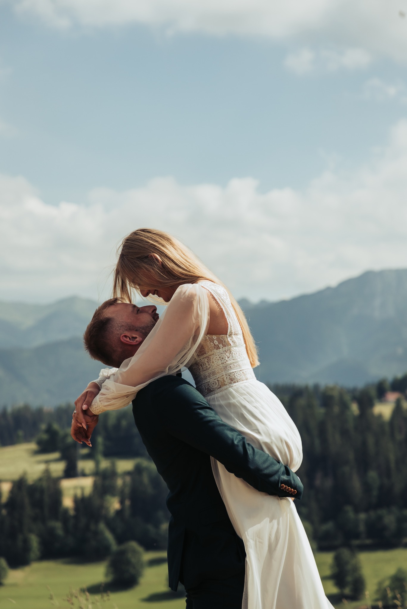 Para młoda w objęciach na tle górskiego krajobrazu. Panna młoda w zwiewnej sukni, pan młody w ciemnym garniturze. Romantyczny kadr z pleneru ślubnego.