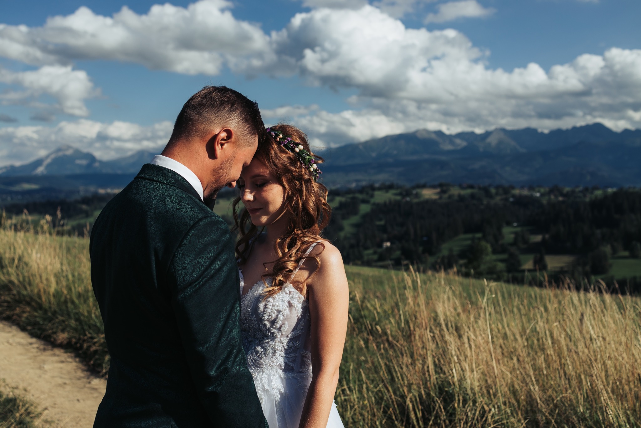Para młoda w objęciach na tle gór i błękitnego nieba z chmurami. Suknia panny młodej zdobiona koronką, pan młody w zielonym garniturze. Romantyczna sceneria plenerowa.