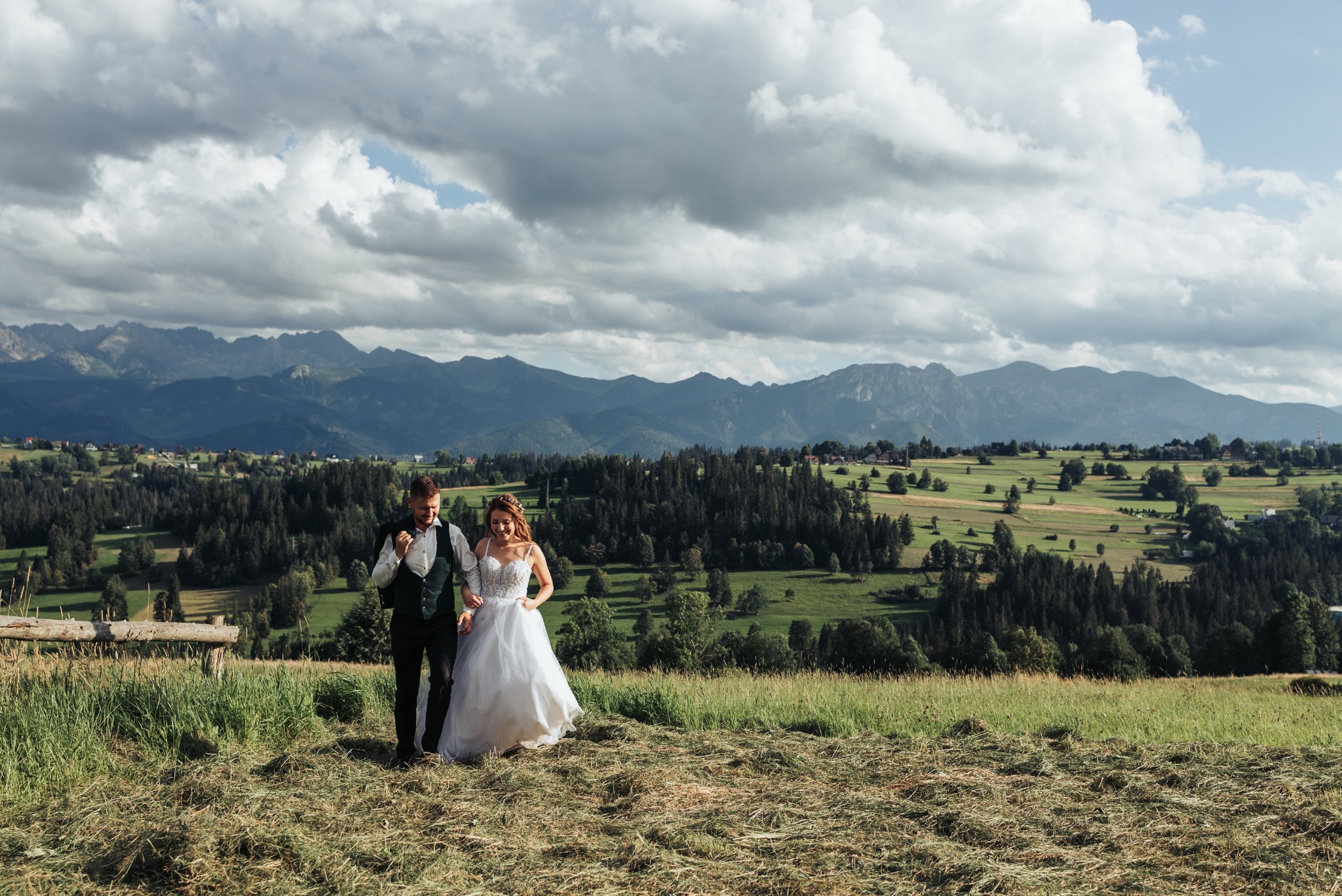 Para młoda spaceruje po łące z widokiem na góry w słoneczny dzień. Panna młoda w białej sukni, pan młody w zielonej kamizelce. Romantyczna sesja ślubna w plenerze.