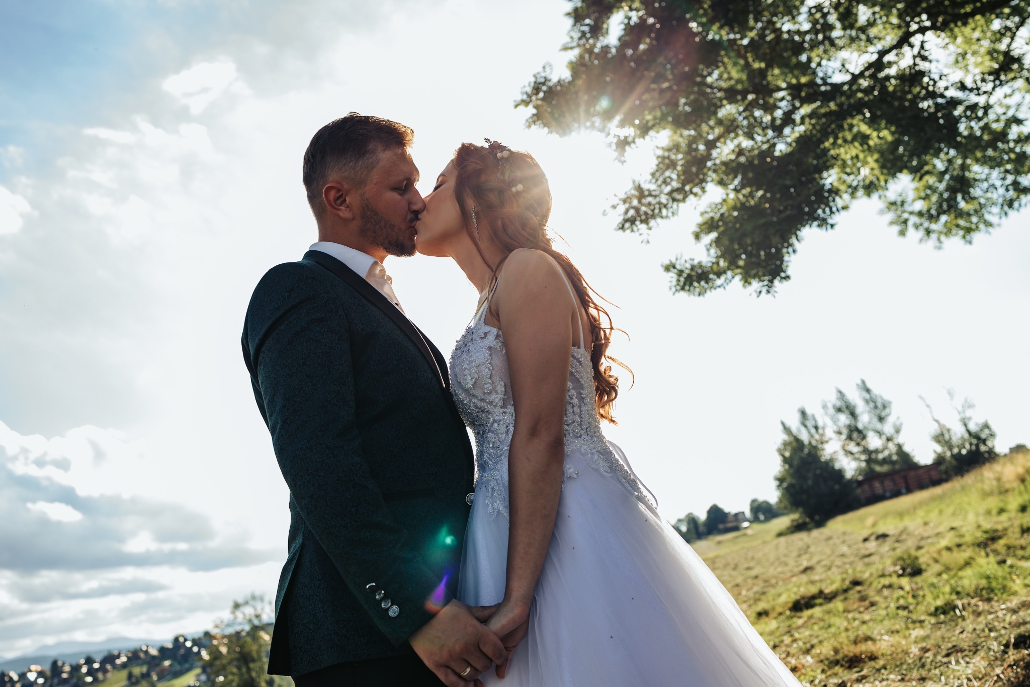 Pocałunek pary młodej w kontraście ze słońcem, on w ciemnozielonym garniturze z wzorem, ona w białej sukni z koronką, trzymają się za ręce na tle łąki.