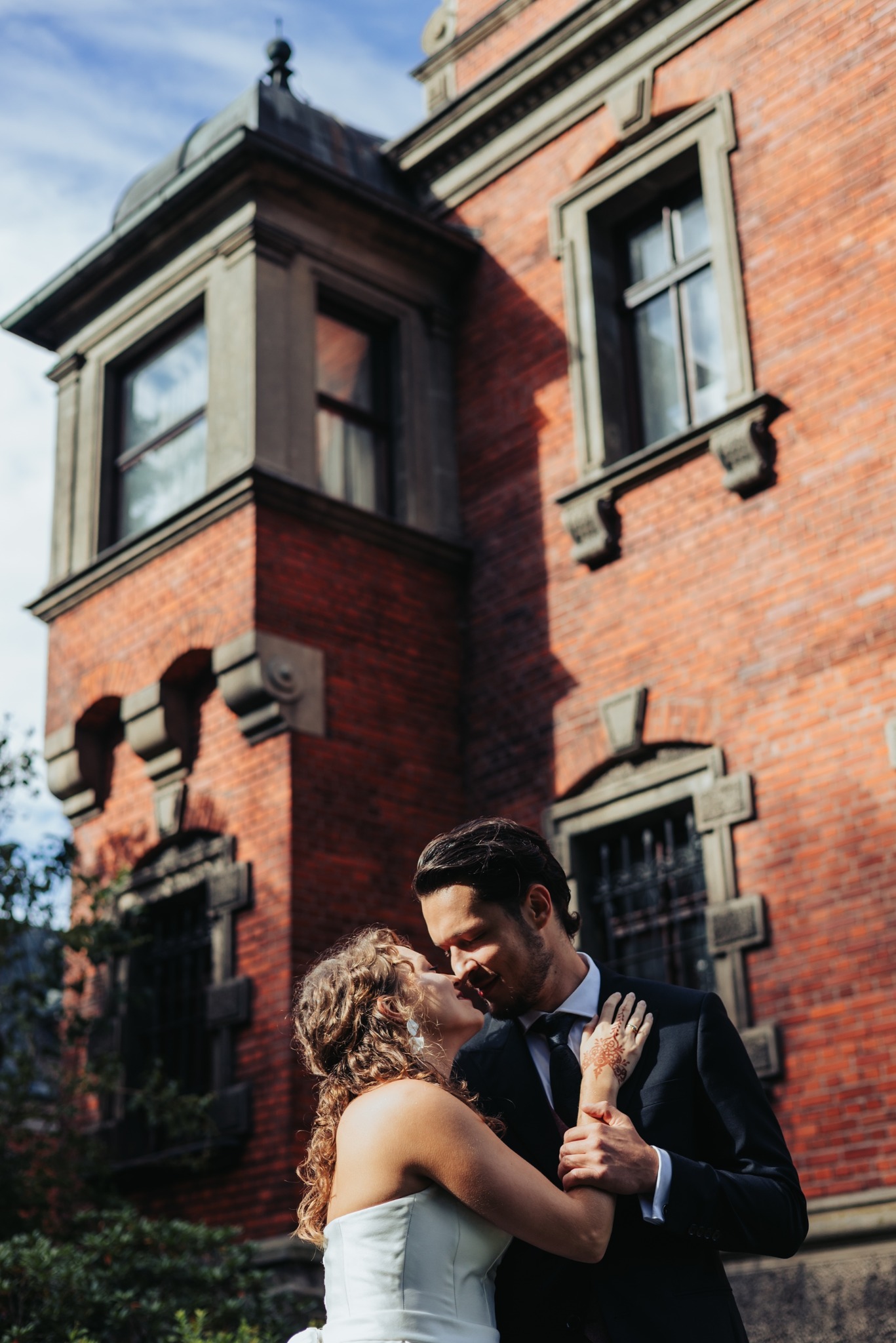 Para młoda w objęciach na tle ceglanej kamienicy z wieżyczką. Panna młoda z henną na dłoni. Ujęcie w ciepłym, naturalnym świetle.