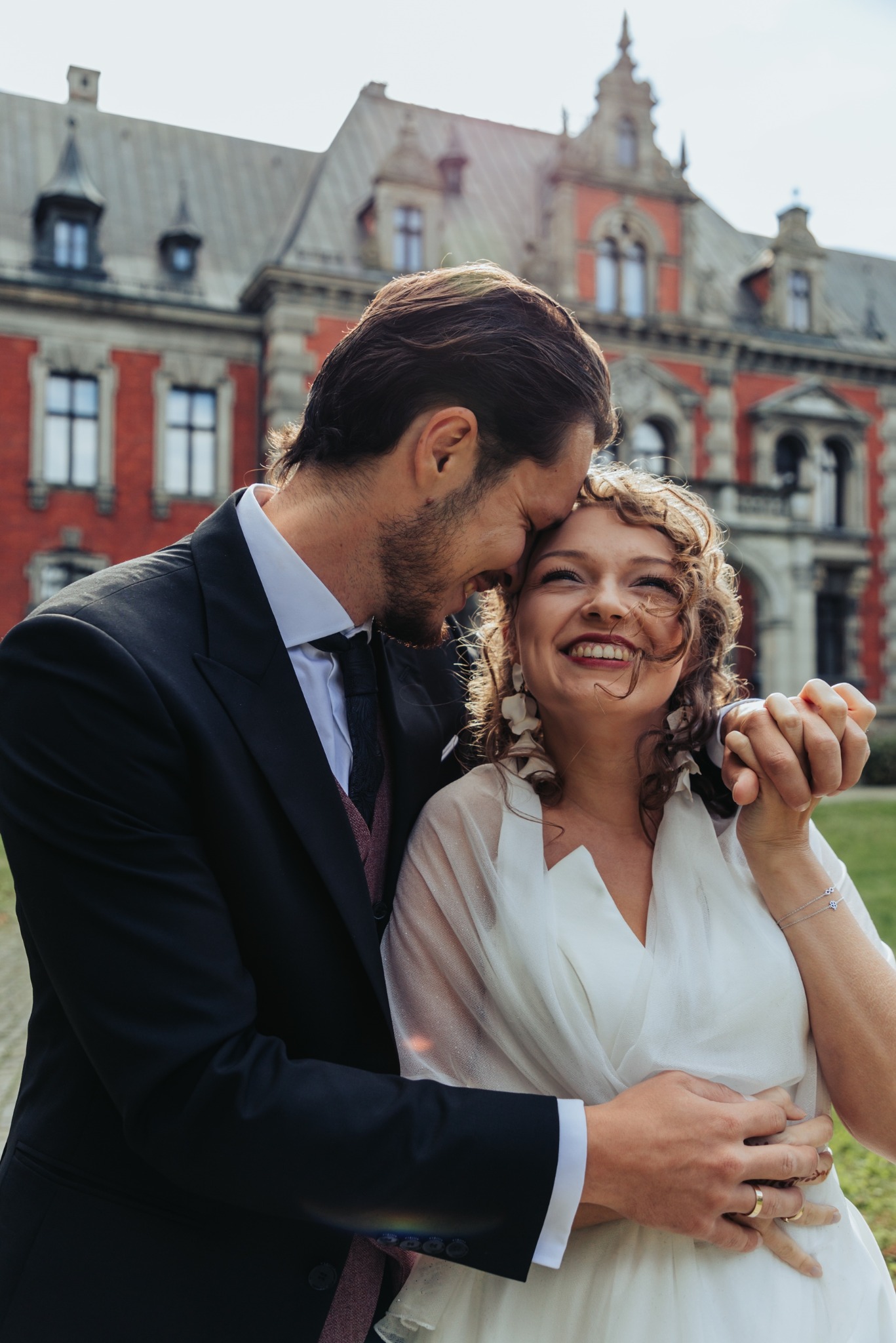 Uśmiechnięta para młoda w objęciach na tle zamku z czerwonej cegły. Pan młody w czarnym garniturze, panna młoda w białej sukni. Romantyczna fotografia ślubna.