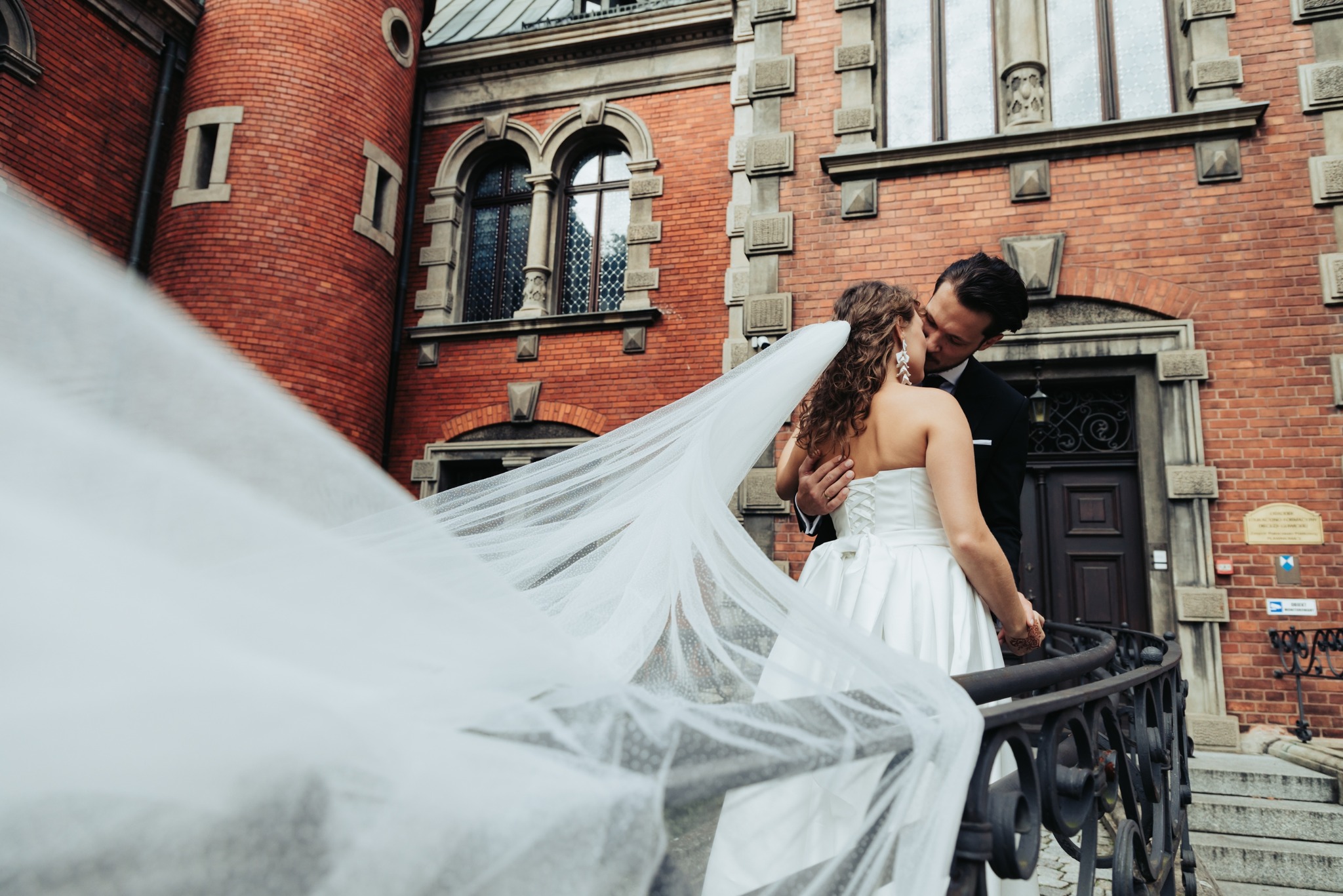 Para młoda w objęciach na tle ceglanej architektury. Długi welon powiewa na wietrze, dodając scenie dynamiki i romantyzmu. Ujęcie z dołu podkreśla monumentalność budynku.