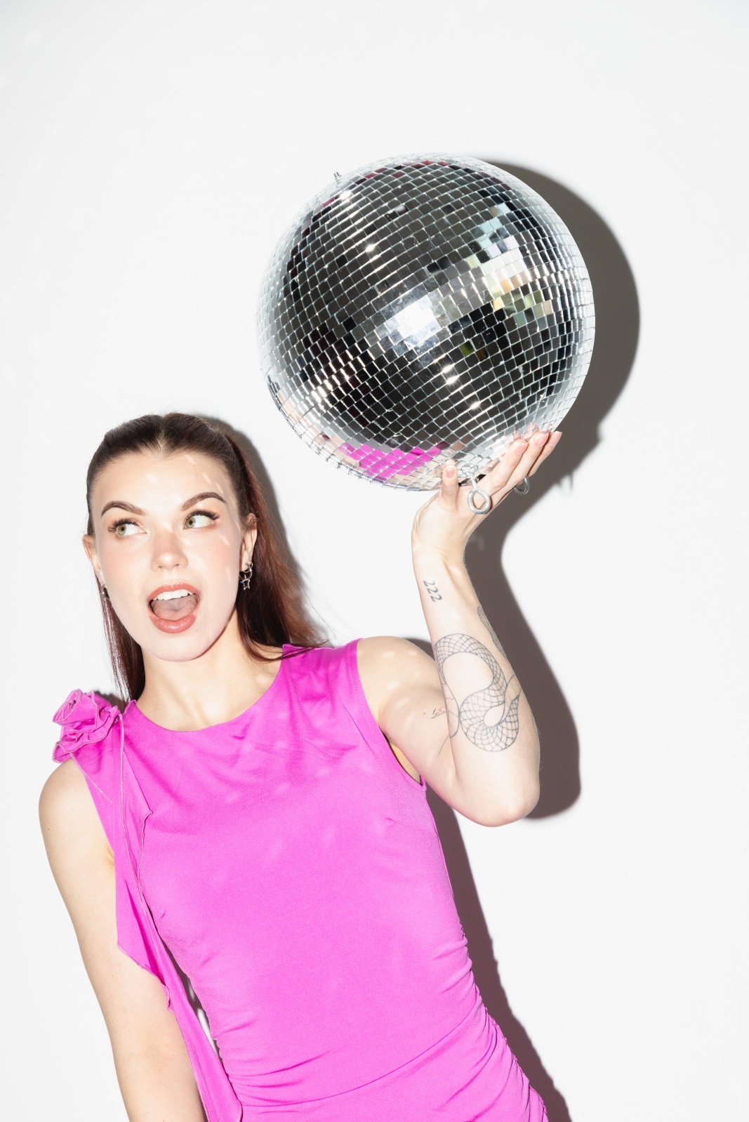 Kobieta w różowej sukience trzymająca błyszczącą kulę dyskotekową, z tatuażem węża na ramieniu, w jasnym oświetleniu i z otwartymi ustami.