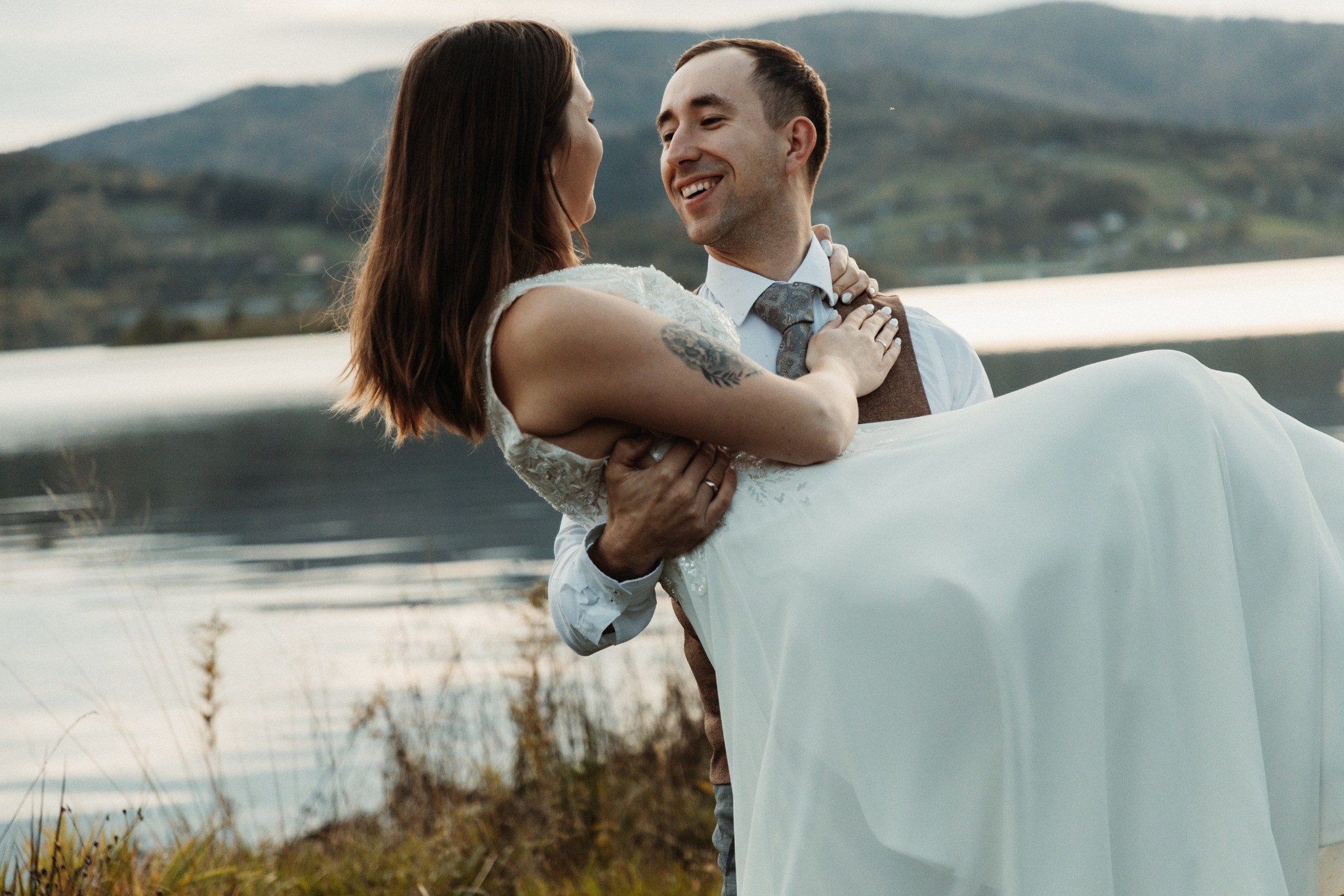 Uśmiechnięty pan młody trzyma pannę młodą w sukni ślubnej na tle jeziora i gór, panna młoda ma tatuaż na ramieniu, oboje patrzą na siebie z czułością.