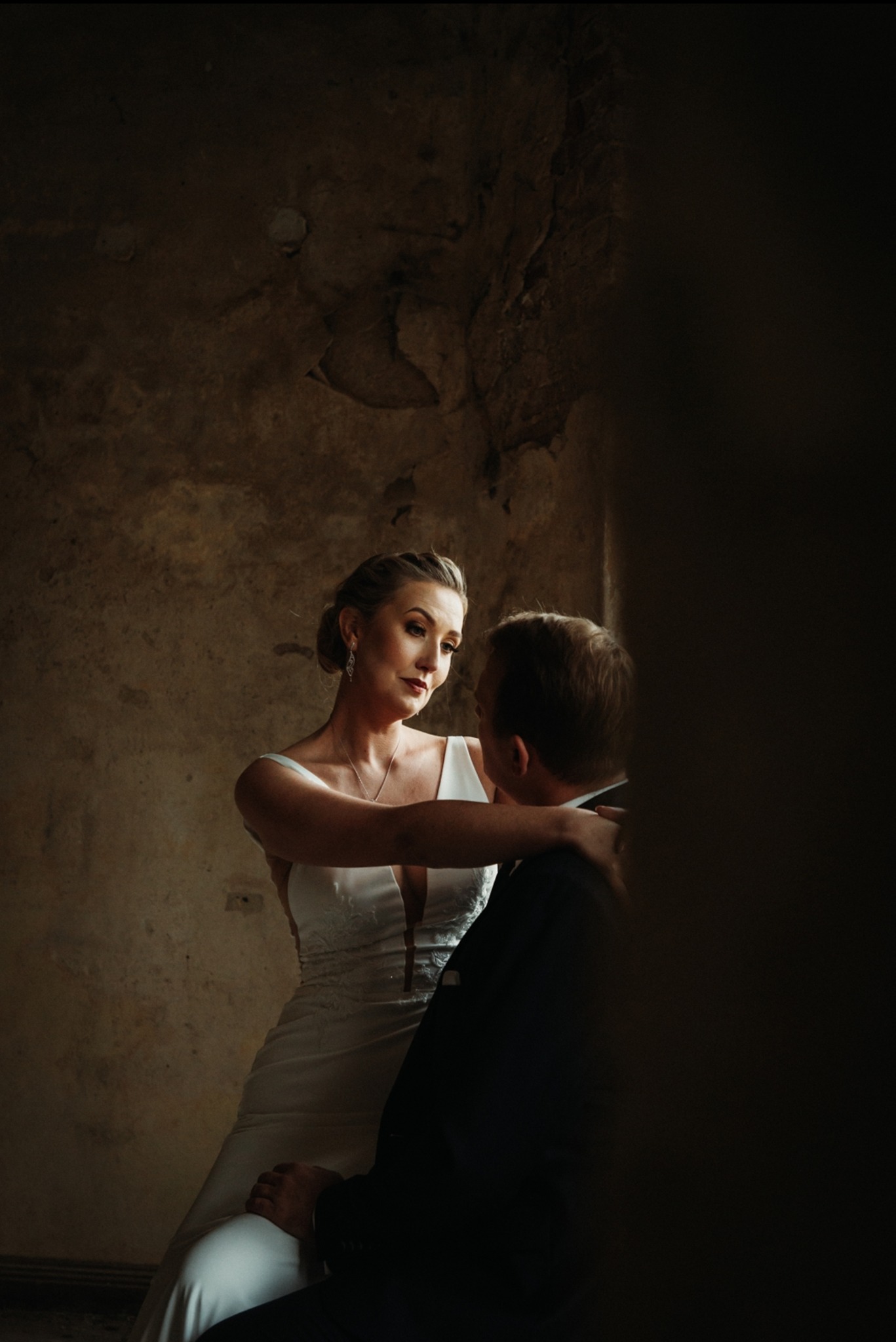 Panna młoda w białej sukni z głębokim dekoltem obejmuje pana młodego w ciemnym garniturze na tle ściany o fakturze starego tynku w ciepłych tonach.