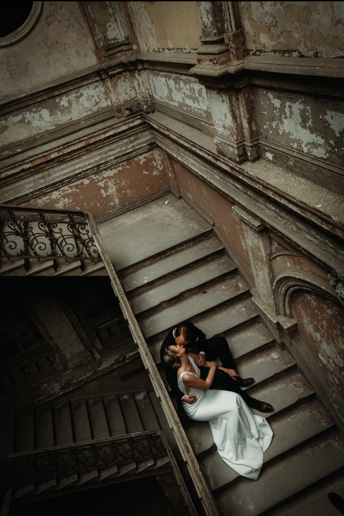 Para młoda w objęciach na schodach starej kamienicy. Ona w białej sukni, on w czarnym garniturze. Ujęcie z góry, widoczne detale zniszczonego wnętrza.