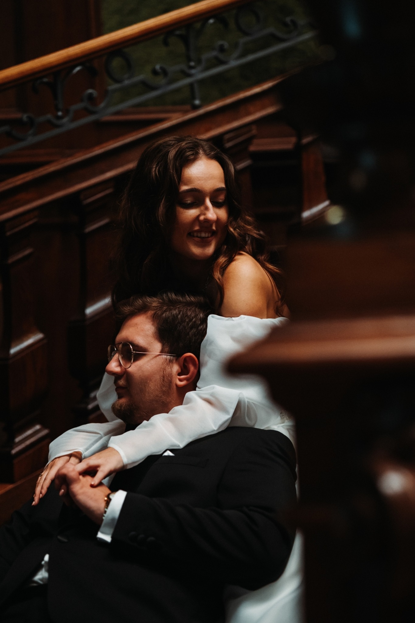 Uśmiechnięta para młoda w objęciach na drewnianych schodach; ona w białej sukni z odkrytymi ramionami, on w czarnym garniturze i okularach.