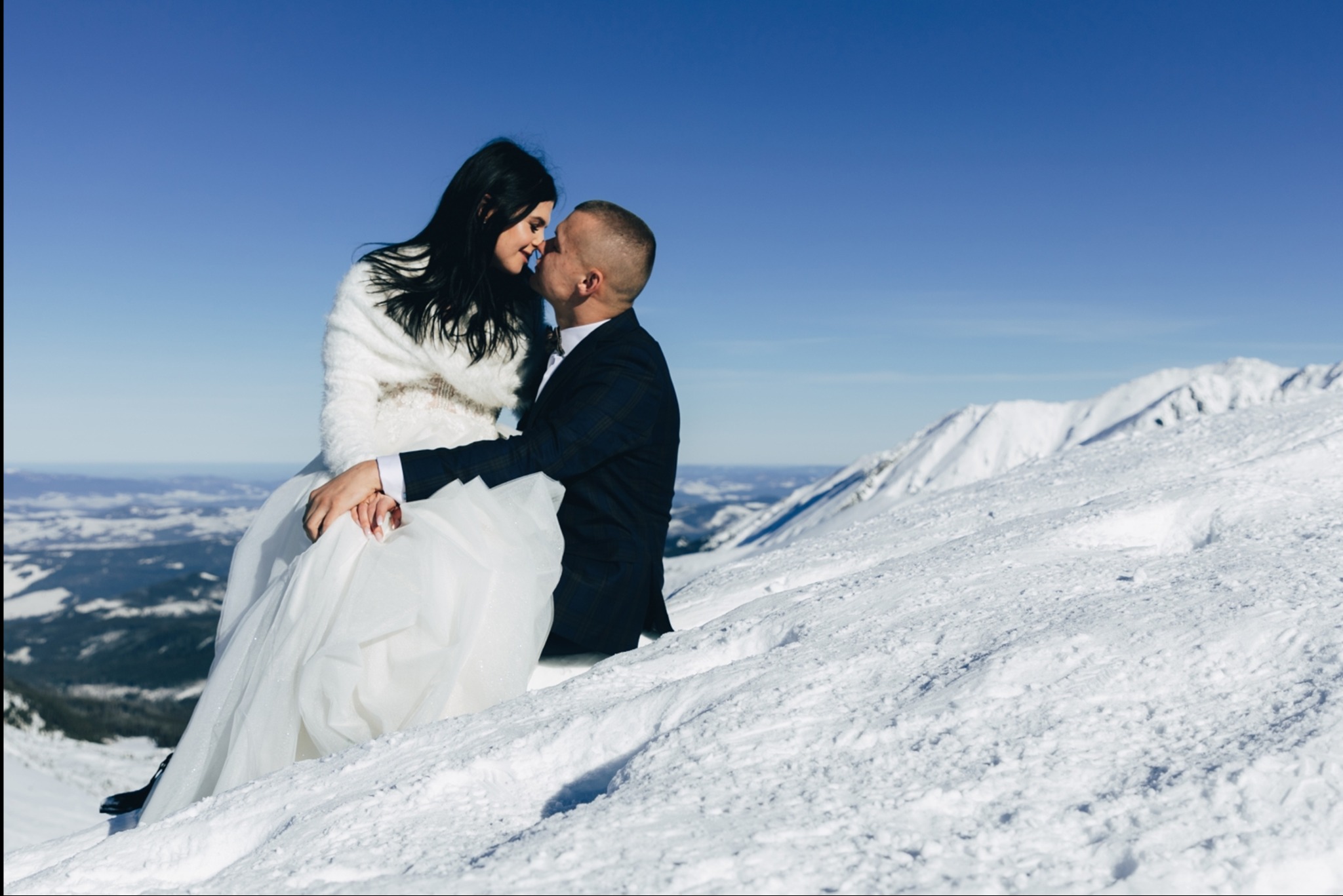 Para młoda w zimowej scenerii górskiej, delikatny pocałunek na tle błękitnego nieba. Panna młoda w białej sukni i futrze, pan młody w granatowym garniturze.