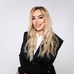 Joanna Płońska - Portret uśmiechniętej blondynki w czarnym żakiecie i białej koszuli na jasnym tle. Profesjonalny wizerunek agenta nieruchomości z delikatnym makijażem.