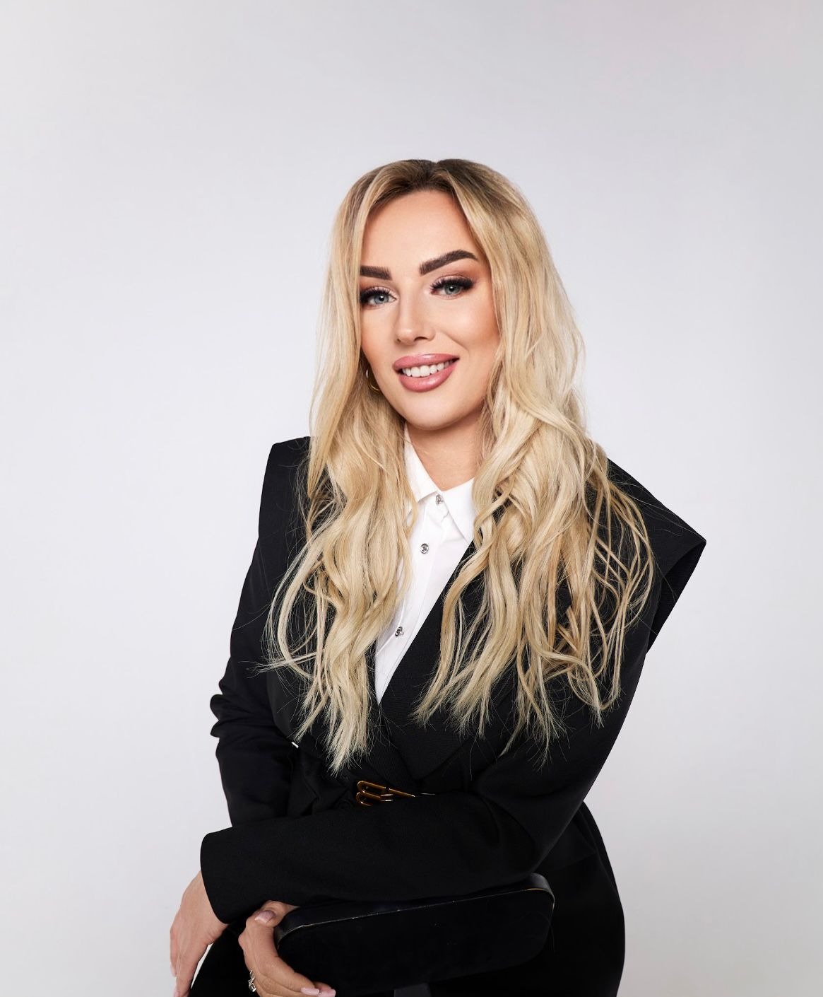 Portret uśmiechniętej blondynki w czarnym żakiecie i białej koszuli na jasnym tle. Profesjonalny wizerunek agenta nieruchomości z delikatnym makijażem.