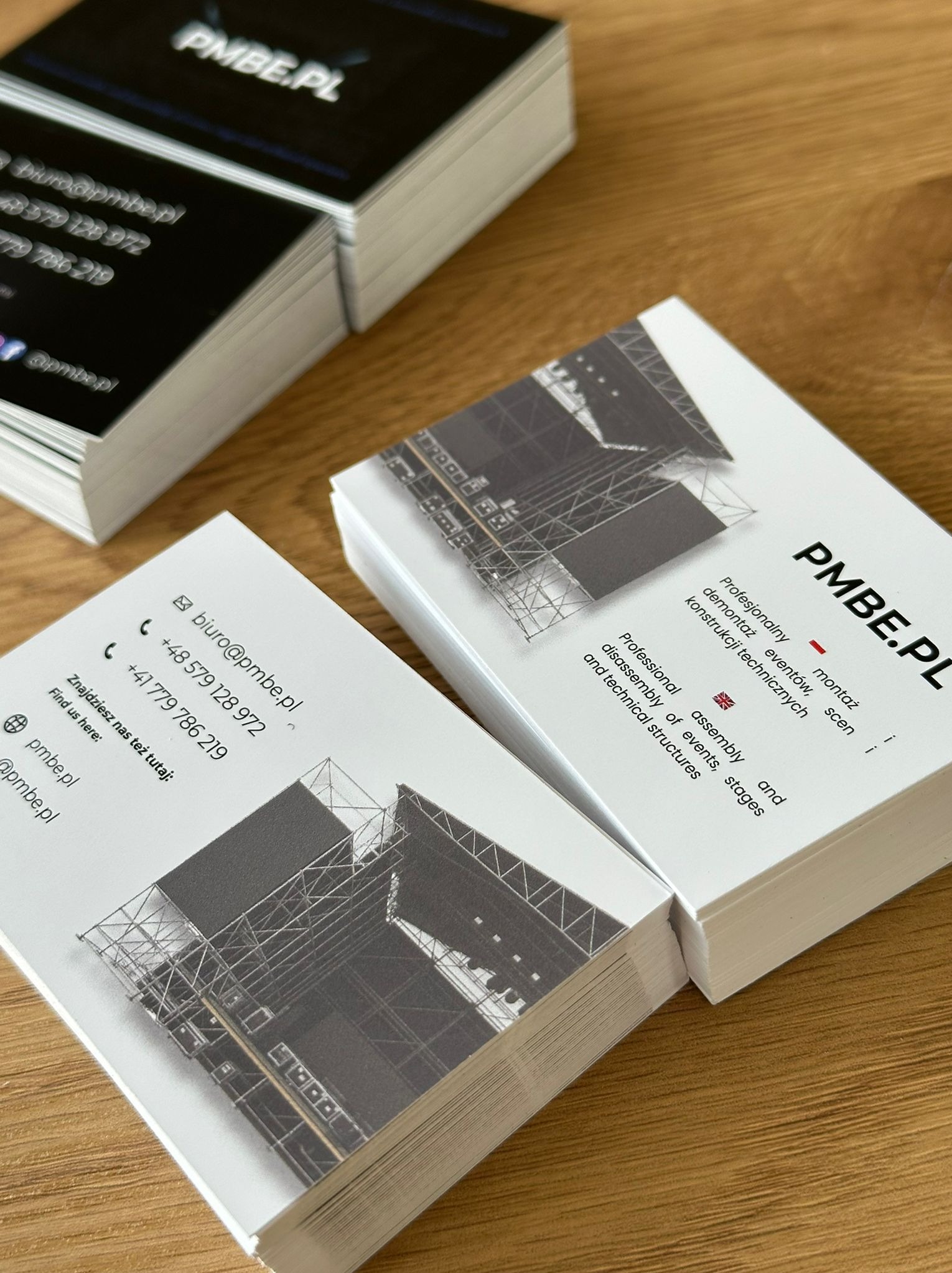 Stos wizytówek firmy PMBE.PL z grafiką sceny i danymi kontaktowymi na drewnianym stole, prezentujący profesjonalny druk i minimalistyczny design.