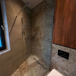 Profi Bud - Nowoczesna łazienka z kabiną prysznicową typu walk-in, wykończona płytkami imitującymi beton i drewnianymi szafkami. Widoczny odpływ liniowy i podtynkowa armatura.