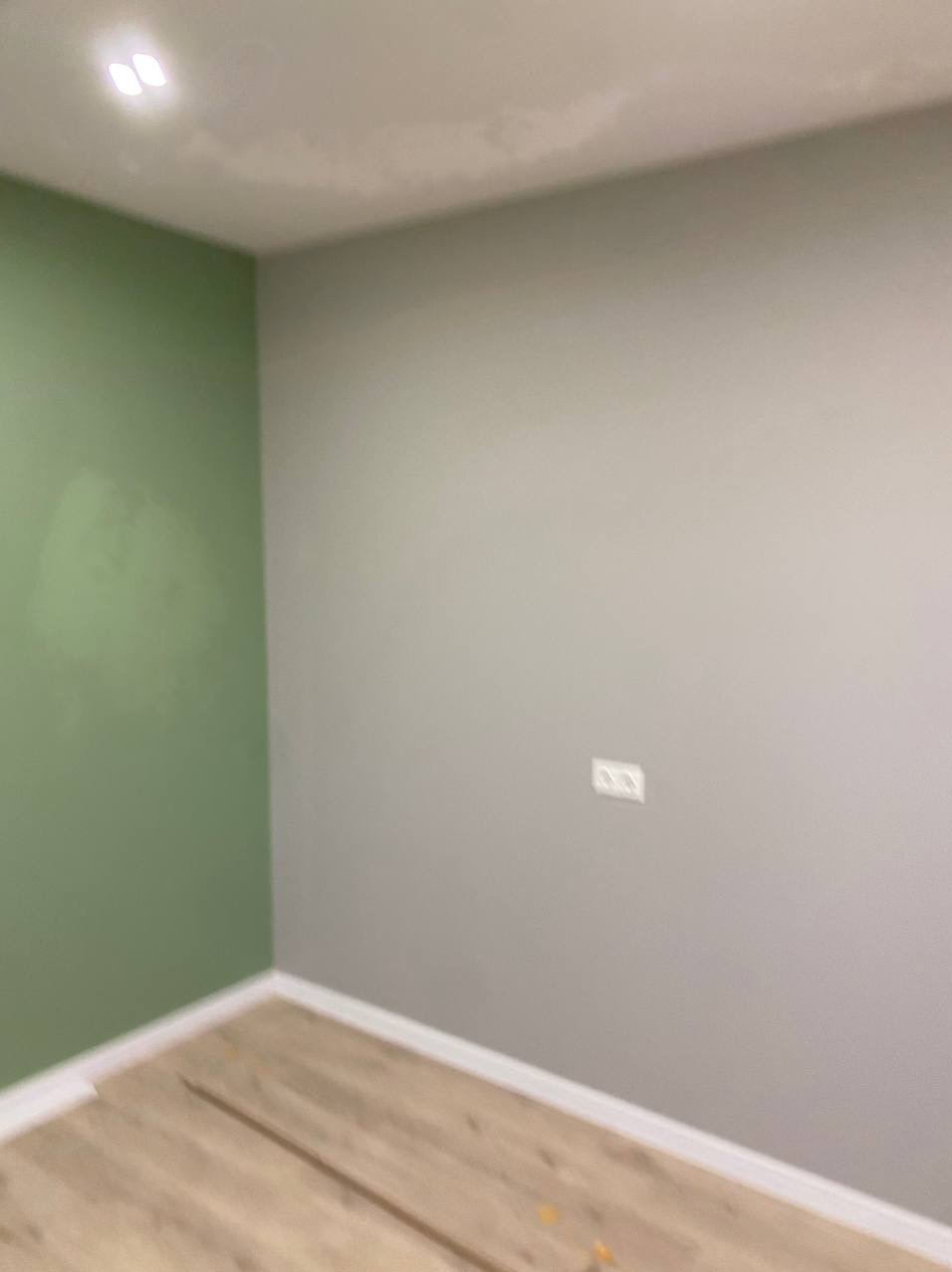 Pomieszczenie po malowaniu: ściana w odcieniach szarości z gniazdkiem, sąsiadująca z zieloną ścianą. Widoczna podłoga z paneli i biały sufit z oświetleniem.