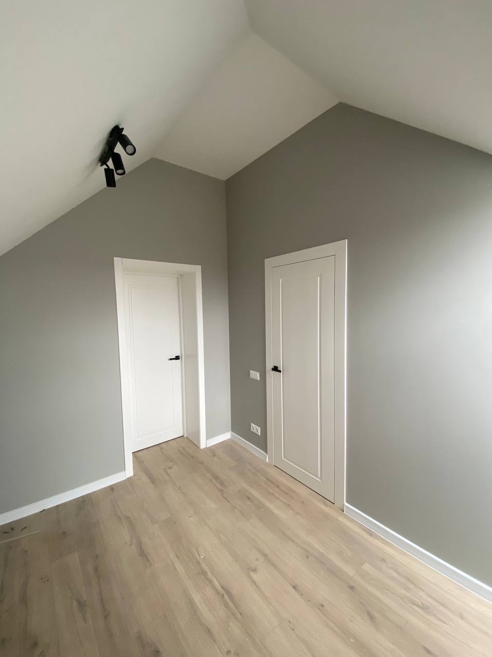 Szara ściana w pokoju na poddaszu z dwoma białymi drzwiami i drewnianą podłogą. Czarny reflektor sufitowy. Minimalistyczny design.