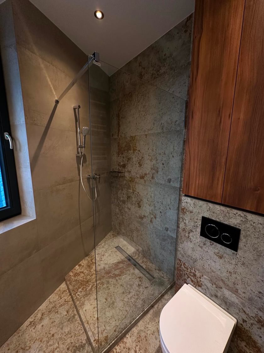 Nowoczesna łazienka z kabiną prysznicową typu walk-in, wykończona płytkami imitującymi beton i drewnianymi szafkami. Widoczny odpływ liniowy i podtynkowa armatura.