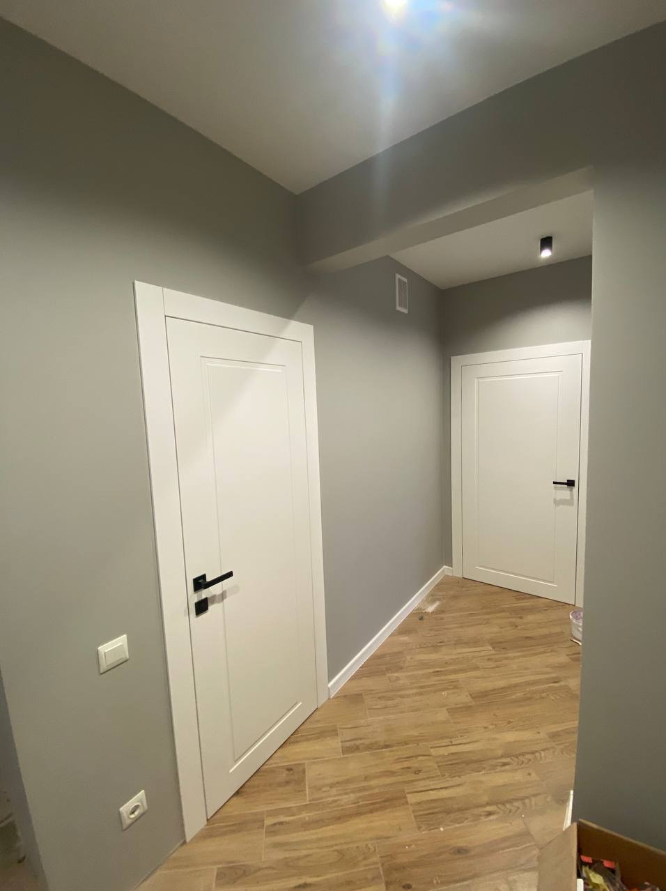 Korytarz po remoncie: szare ściany, drewnopodobna podłoga i dwie białe drzwi z czarnymi klamkami. Minimalistyczne wykończenie, nowoczesny styl.