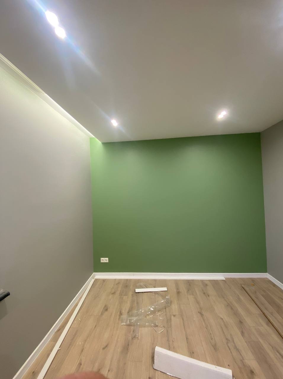 Świeżo pomalowane wnętrze: zielona ściana kontrastuje z szarą, drewniana podłoga. Widoczne listwy przypodłogowe i oświetlenie sufitowe.