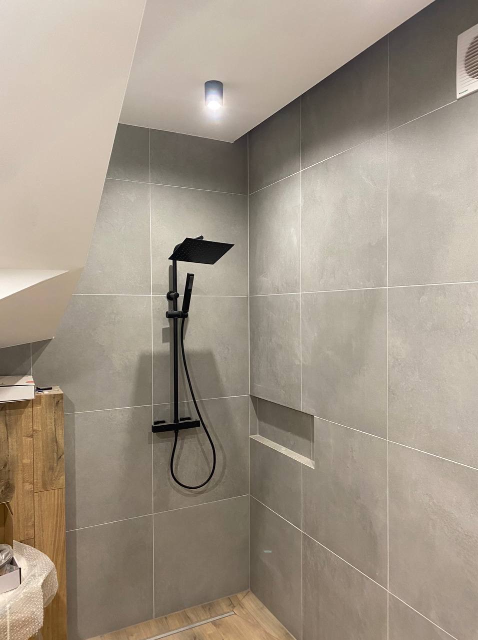 Wykończona łazienka na poddaszu: szare płytki wielkoformatowe, czarna armatura natryskowa, wnęka w ścianie. Minimalistyczny design, nowoczesne oświetlenie.