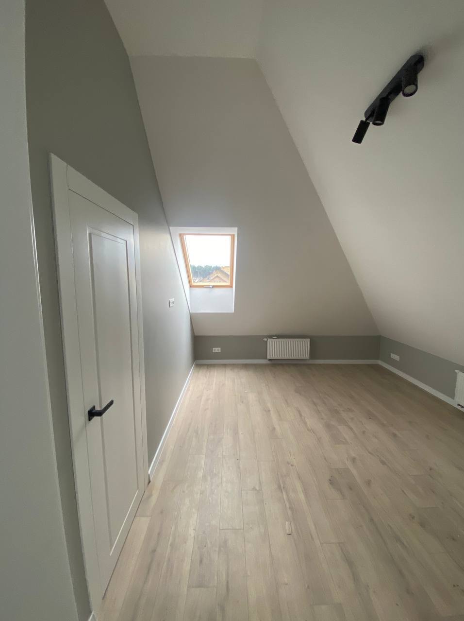 Jasne poddasze po remoncie: skośne ściany, drewniana podłoga, białe drzwi z czarną klamką i okno dachowe. W tle grzejnik i szara lamperia.