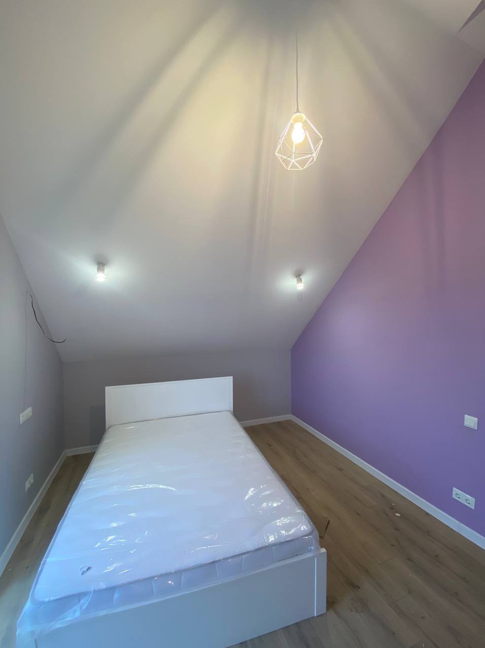 Pokój na poddaszu po remoncie: biały sufit, fioletowa ściana, drewniana podłoga, białe łóżko z folią ochronną i nowoczesna lampa wisząca.