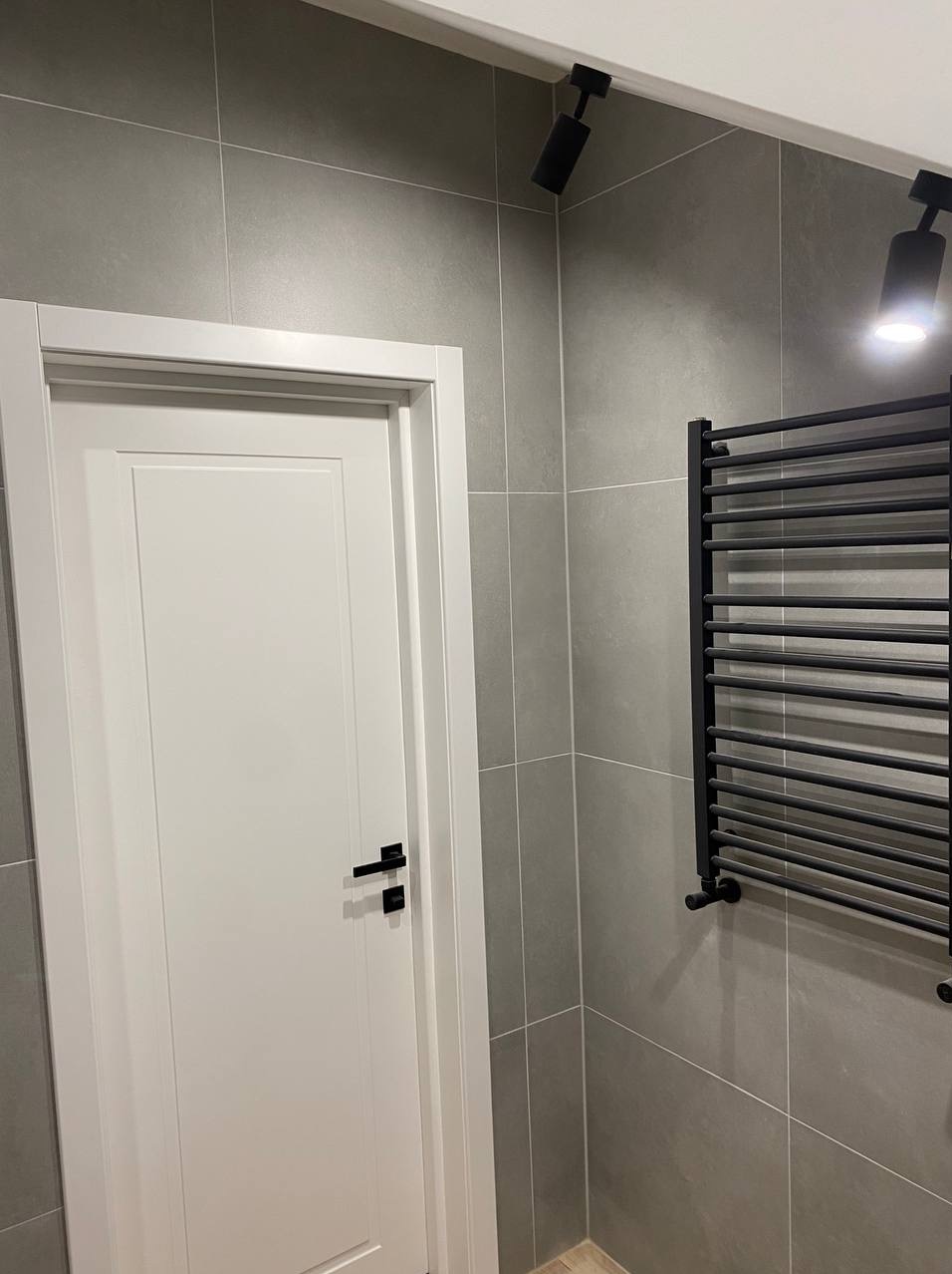 Wykończona łazienka z szarymi płytkami, białą stolarką drzwiową i czarnym grzejnikiem drabinkowym. Minimalistyczne oświetlenie punktowe.