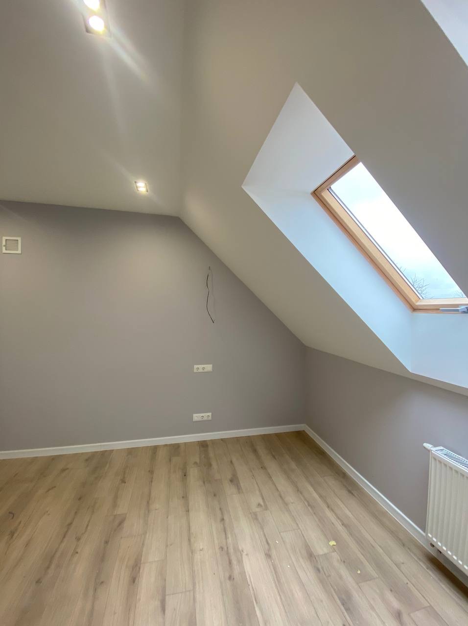 Pomalowane na szaro poddasze ze skośnym oknem dachowym i panelami podłogowymi. Widoczne gniazdka elektryczne, listwy przypodłogowe i grzejnik. Ujęcie z dołu.