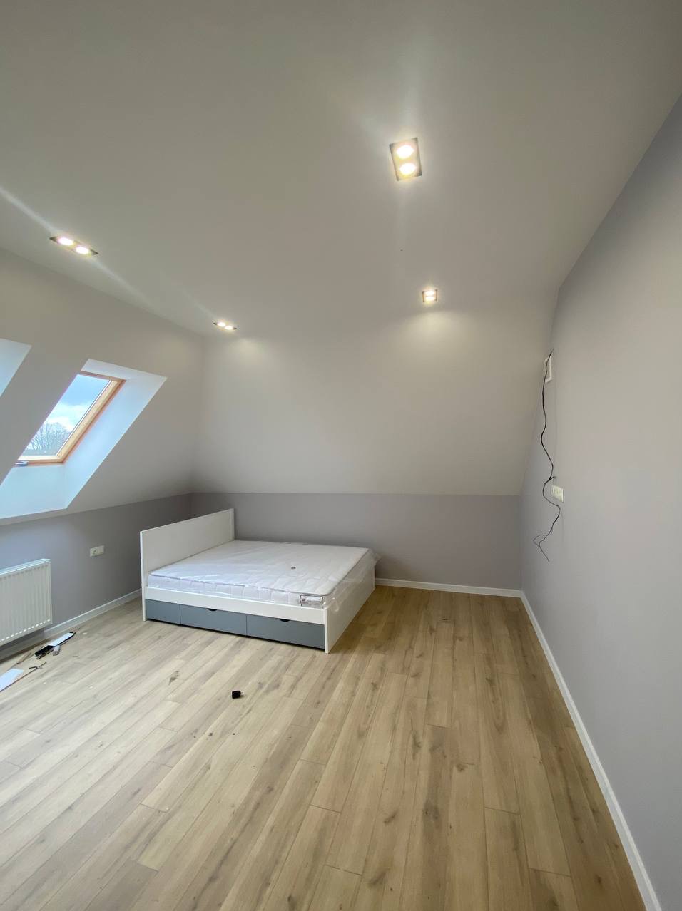 Jasne poddasze po remoncie: drewniana podłoga, szara lamperia, białe ściany, okno dachowe i łóżko z szufladami. Minimalistyczne oświetlenie sufitowe.
