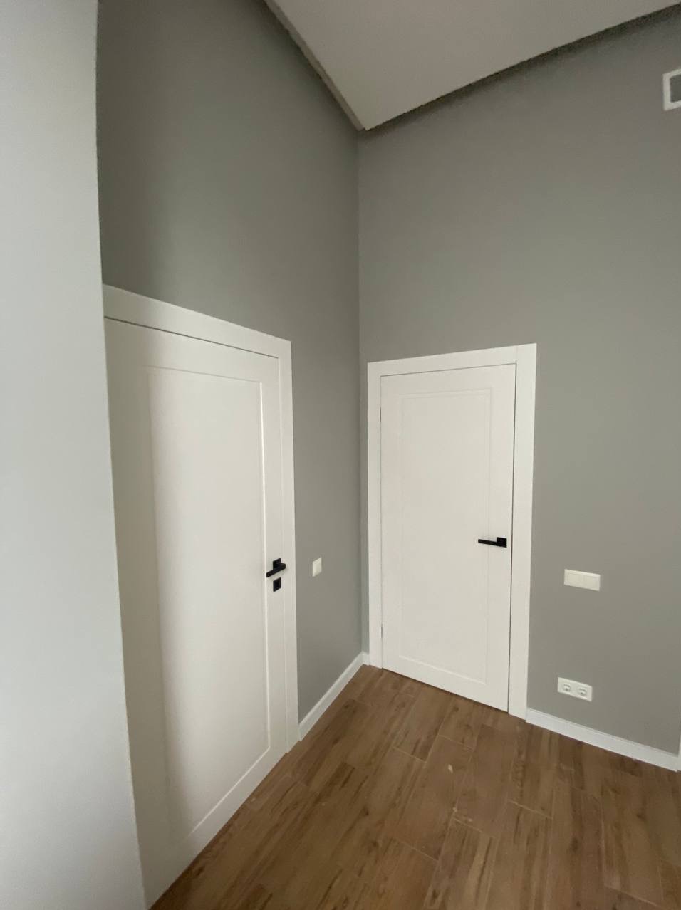 Szare ściany i dwie białe, minimalistyczne drzwi z czarnymi klamkami. Podłoga wyłożona brązowymi panelami imitującymi drewno. Nowoczesne wykończenie wnętrza.