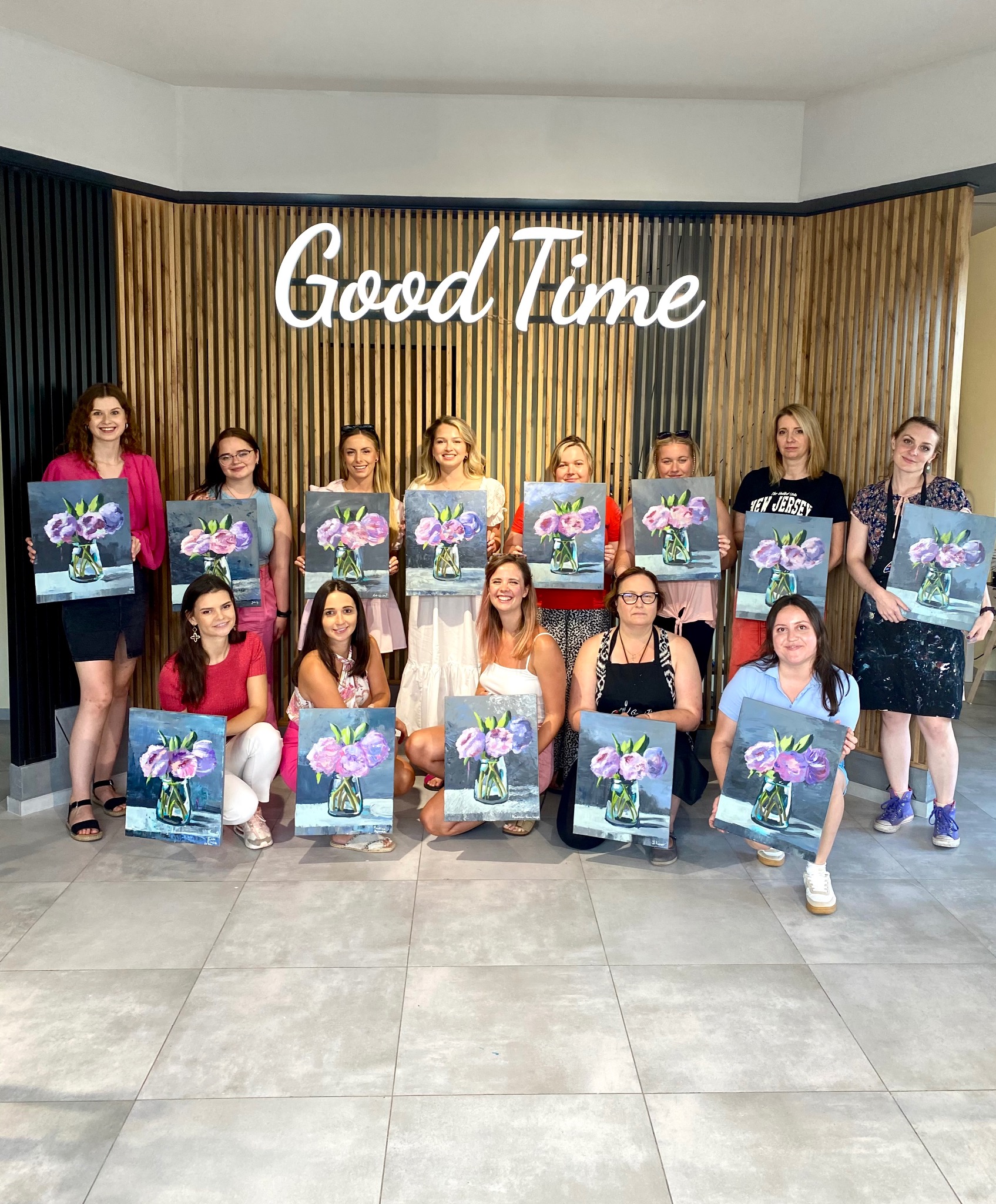 Grupa kobiet prezentuje samodzielnie namalowane obrazy z motywem kwiatów w wazonie, na tle ściany z napisem 'Good Time'.