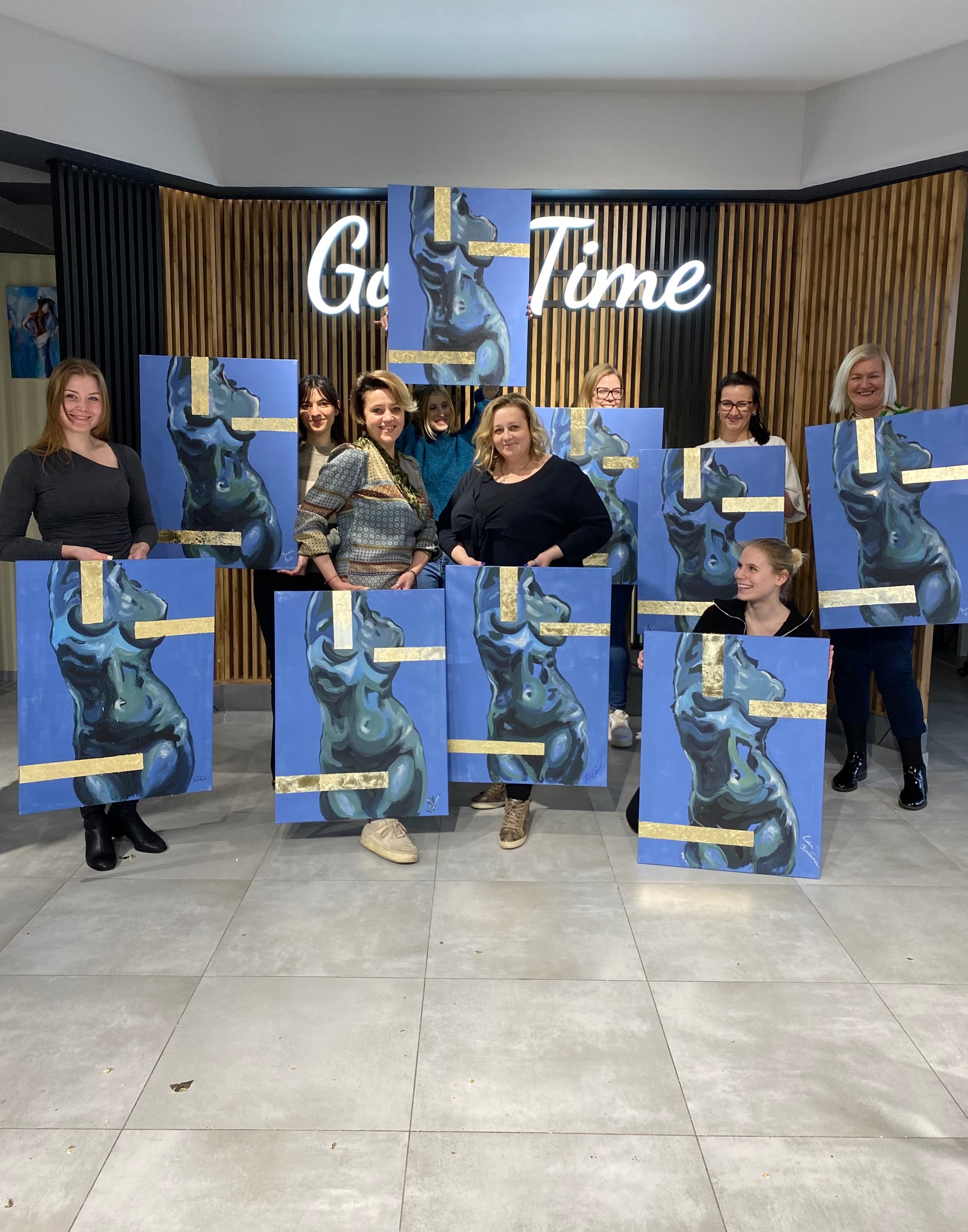 Grupa kobiet prezentuje obrazy ciał w odcieniach błękitu z elementami złota, wykonane prawdopodobnie podczas warsztatów artystycznych. W tle napis 'Good Time'.